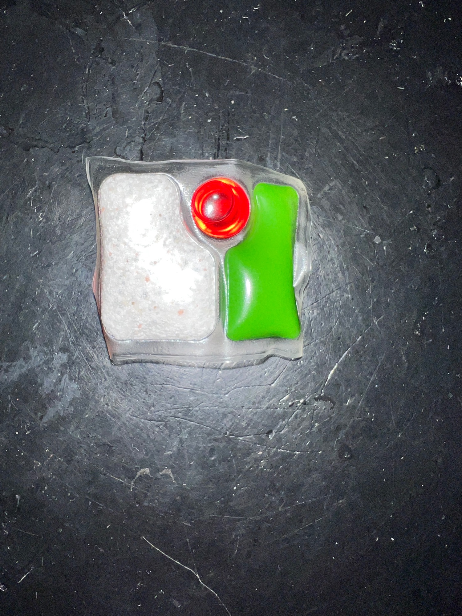 Une pastille de détergent pour lave-vaisselle, triple action, effet retardé, avec une poudre blanche, un liquide rouge et un gel vert.